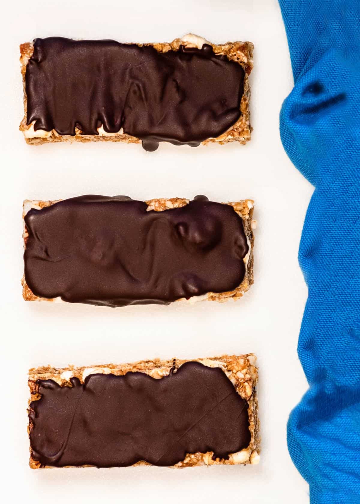 3 chocolate covered peanut butter s'mores granola bars on a white counter with a blue towel next to it.