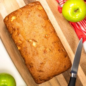 Fresh baked loaf of apple cinnamon bread on a cutting board with a knife and green apples next to it.
