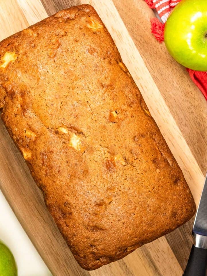 Fresh baked loaf of apple cinnamon bread on a cutting board with a knife and green apples next to it.
