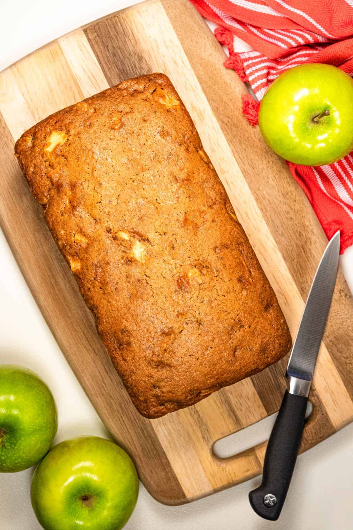 Freshly baked loaf of apple cinnamon bread sitting on a cutting board. Laying around the cutting board are green apples, a knife, and a red towel.
