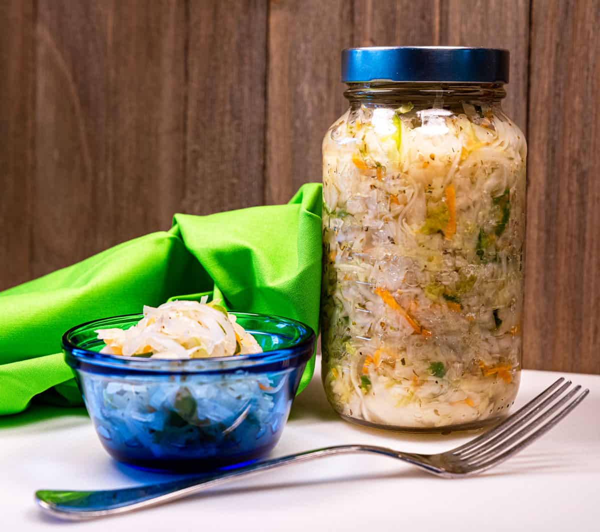 A small blue dish of salvadoran curtido slaw with a fork and mason jar of it next to it.