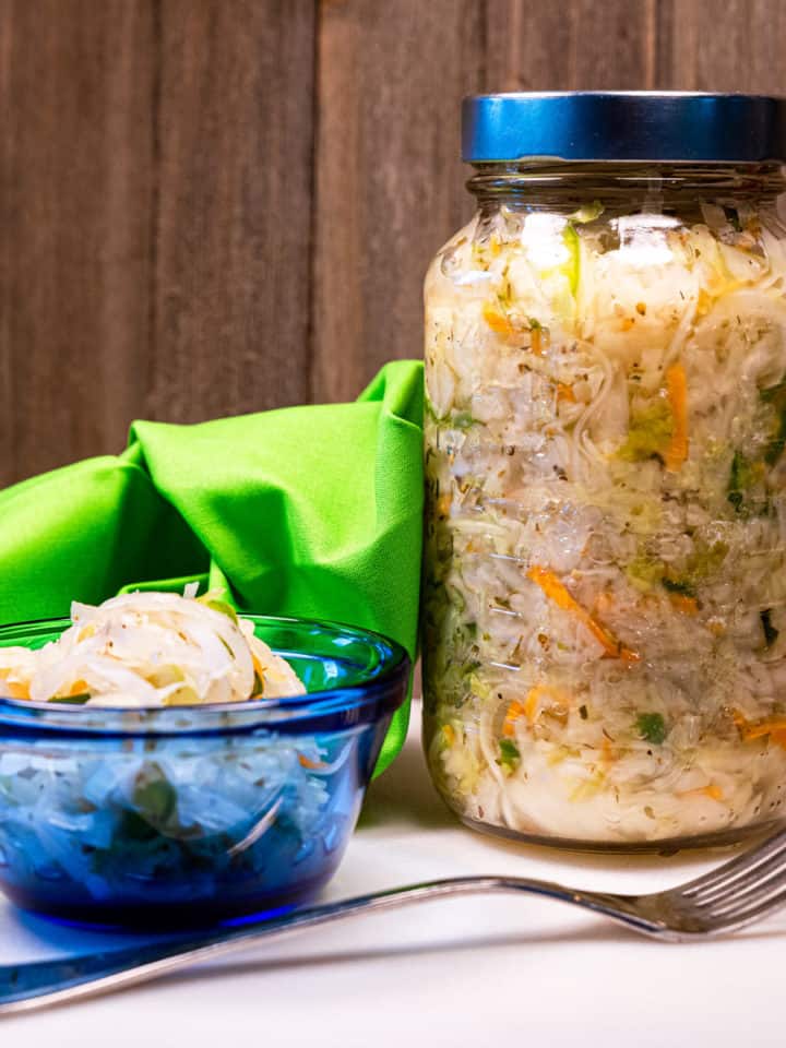 A small blue dish of salvadoran curtido slaw with a fork and mason jar of it next to it.