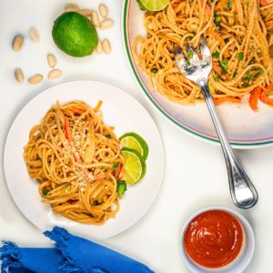 Overhead view of a plate of peanut noodles next to a big serving dish of the noodles. There are peanuts around the plate and lime.