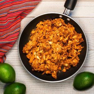 A skillet of jackfruit carnitas on a white wood table with a red towel and limes.