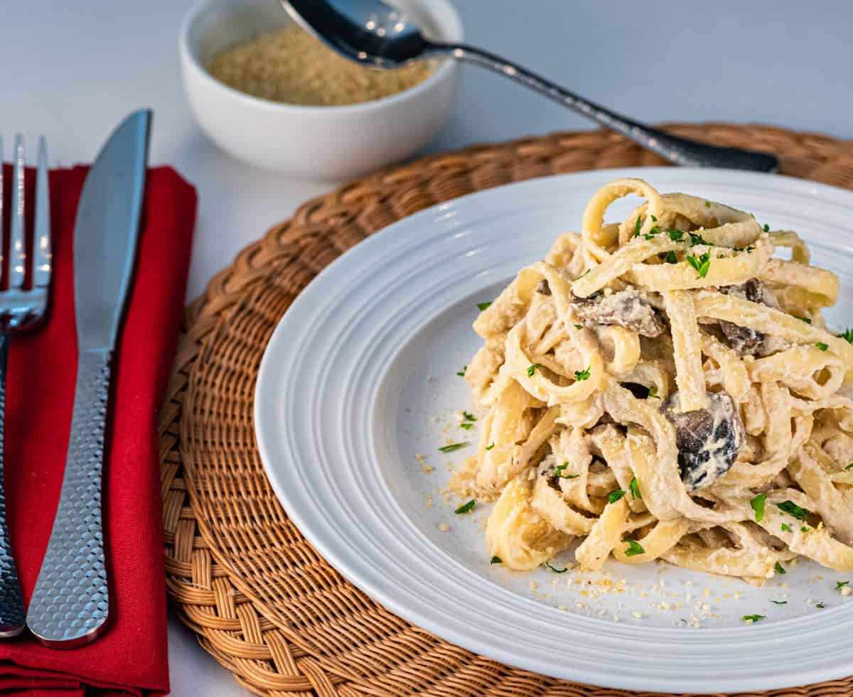 A plate of vegan mushroom fettuccine alfredo on a white plate with vegan parmesan cheese in a small bowl behind the plate. Next to it is a red cloth napkin with a fork and knife.