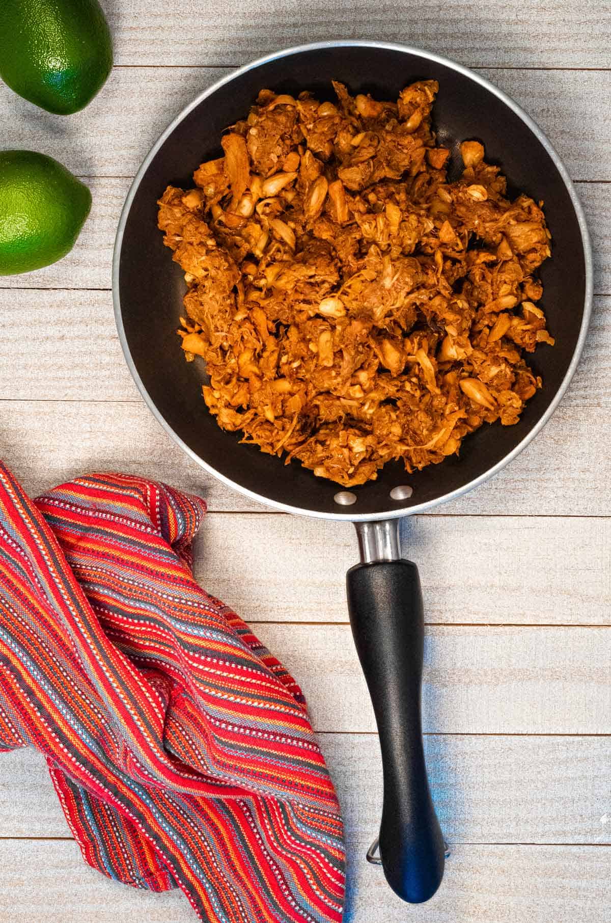 A skillet of jackfruit carnitas on a white wood table with a red towel and limes.
