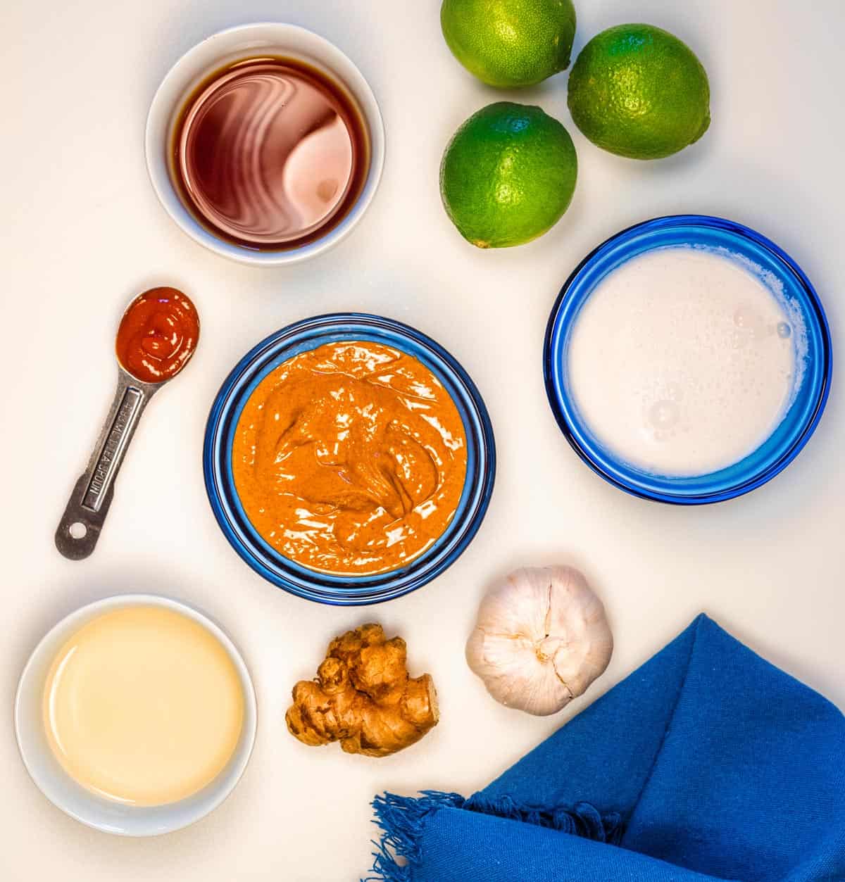 A dish of peanut butter, dish or coconut milk, piece of ginger, clove of garlic, three limes and teaspoon of hot sauce all ready to make peanut sauce.