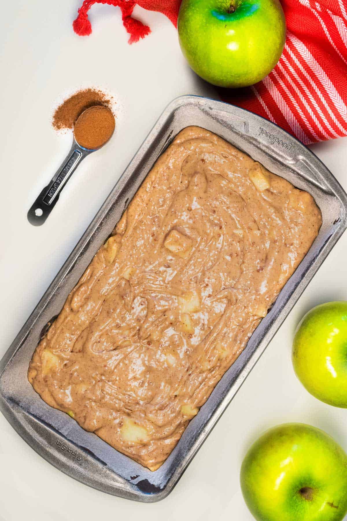 A pan of apple cinnamon loaf batter ready to be baked with a teaspoon of cinnamon and green apples next to it.