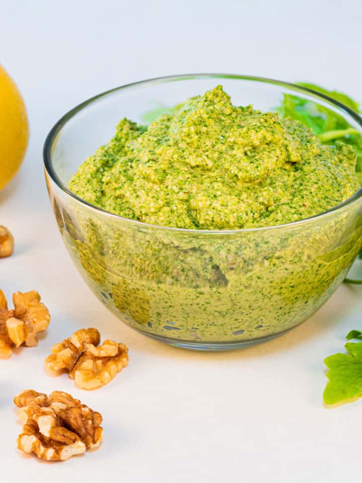 A bowl of arugula walnut pesto sauce with a lemon in the background and some walnuts scattered around.