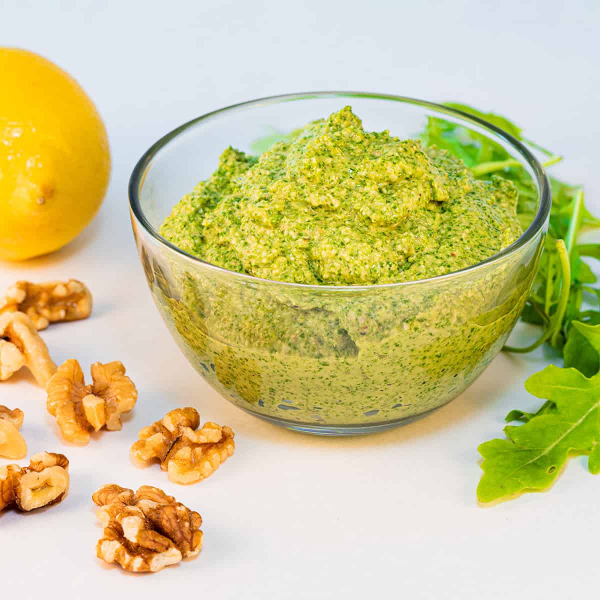 A bowl of arugula walnut pesto sauce with a lemon in the background and some walnuts scattered around.