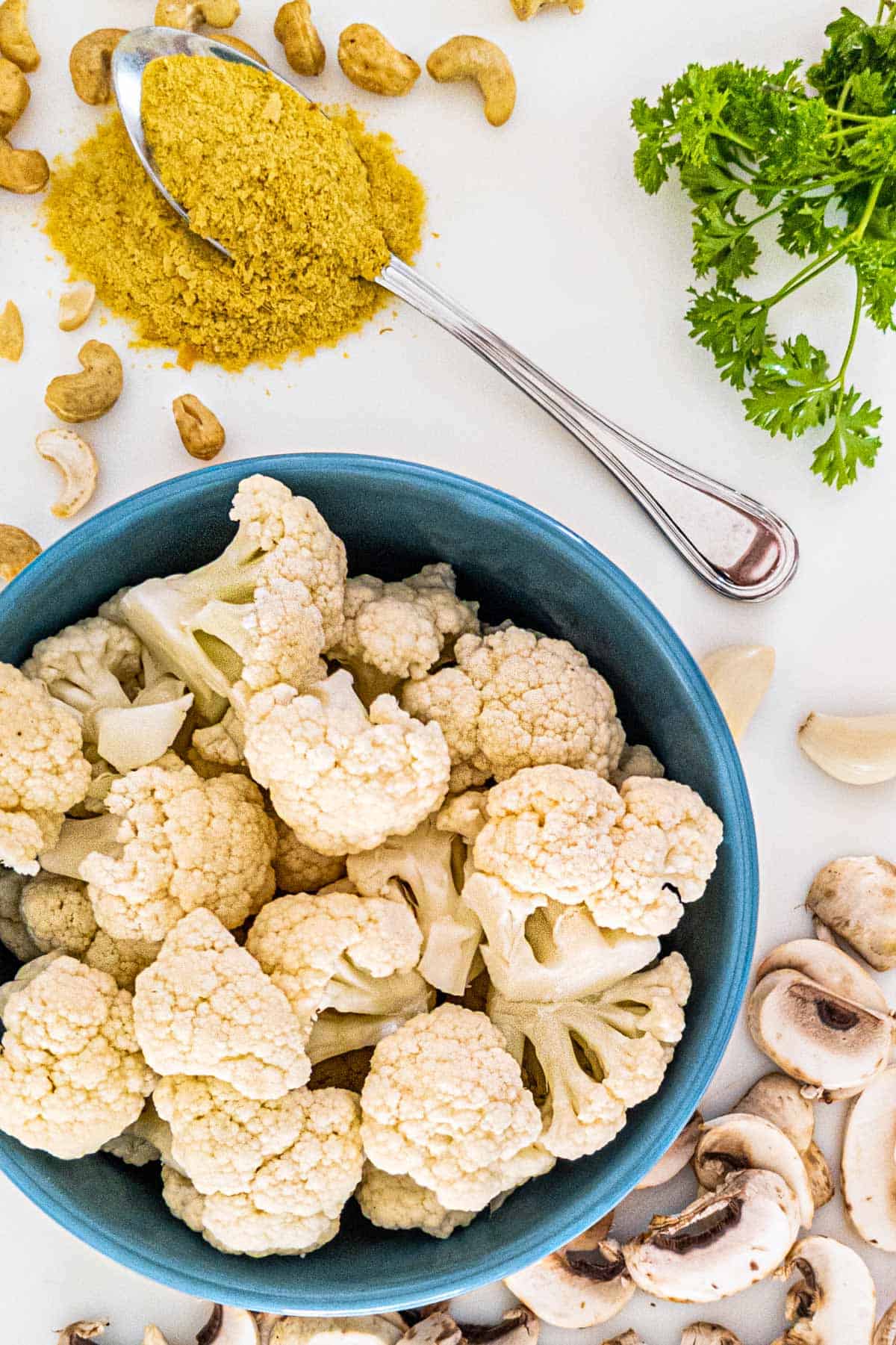 Cauliflower, mushroom, cashews, garlic cloves, parsley, and a spoon of nutritional yeast on a white board.