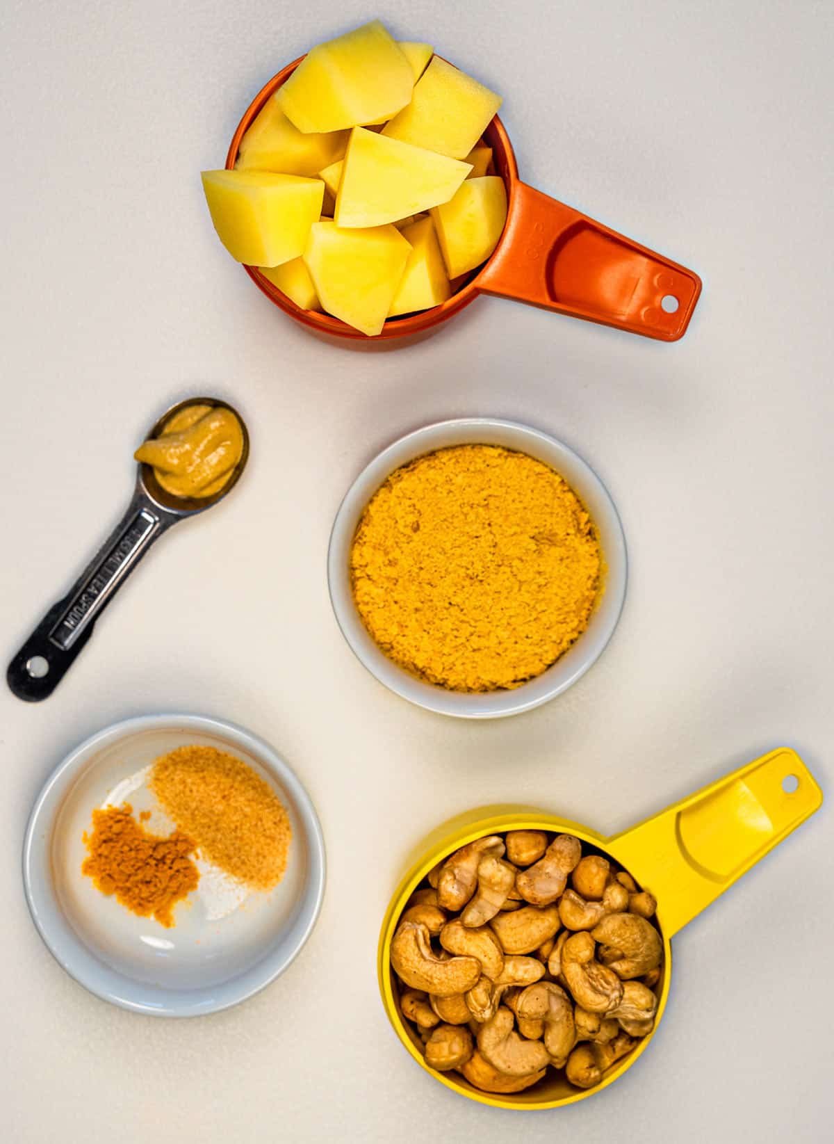 Orange measuring cup of cubed potatoes, yellow measuring cup of cashews, white bowl of nutritional yeast, measuring spoon of mustard, and white bowl of spices.