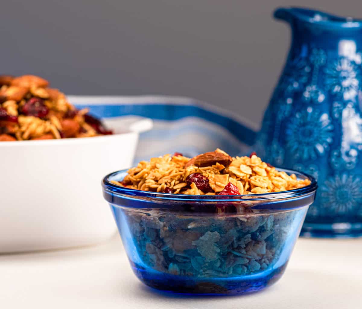 Bowl of vanilla cranberry coconut granola with almonds and cranberries in a blue bowl with a blue pitcher in the background.