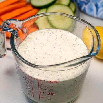 Dish of vegan ranch dressing with other vegetables surrounding the dish like carrot sticks, a lemon, and some cucumber slices.