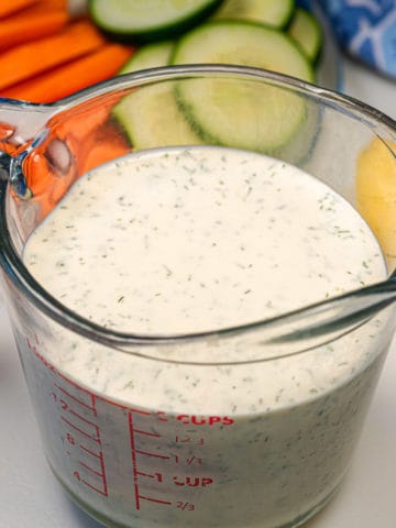 Dish of vegan ranch dressing with other vegetables surrounding the dish like carrot sticks, a lemon, and some cucumber slices.