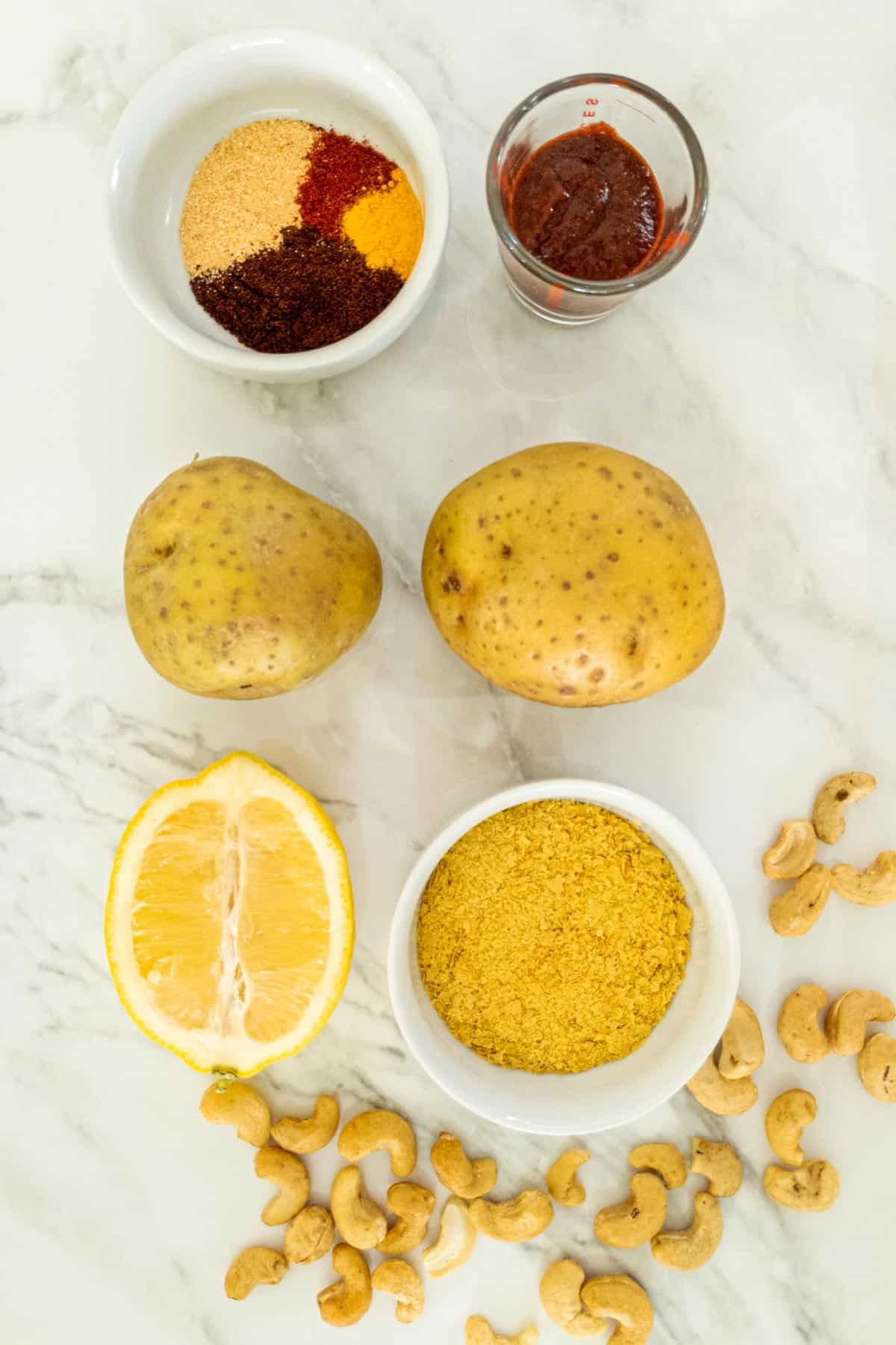 Two potatoes, dish of spices, dish or nutritional yeast, half a lemon, and scattered cashews on a marble counter.
