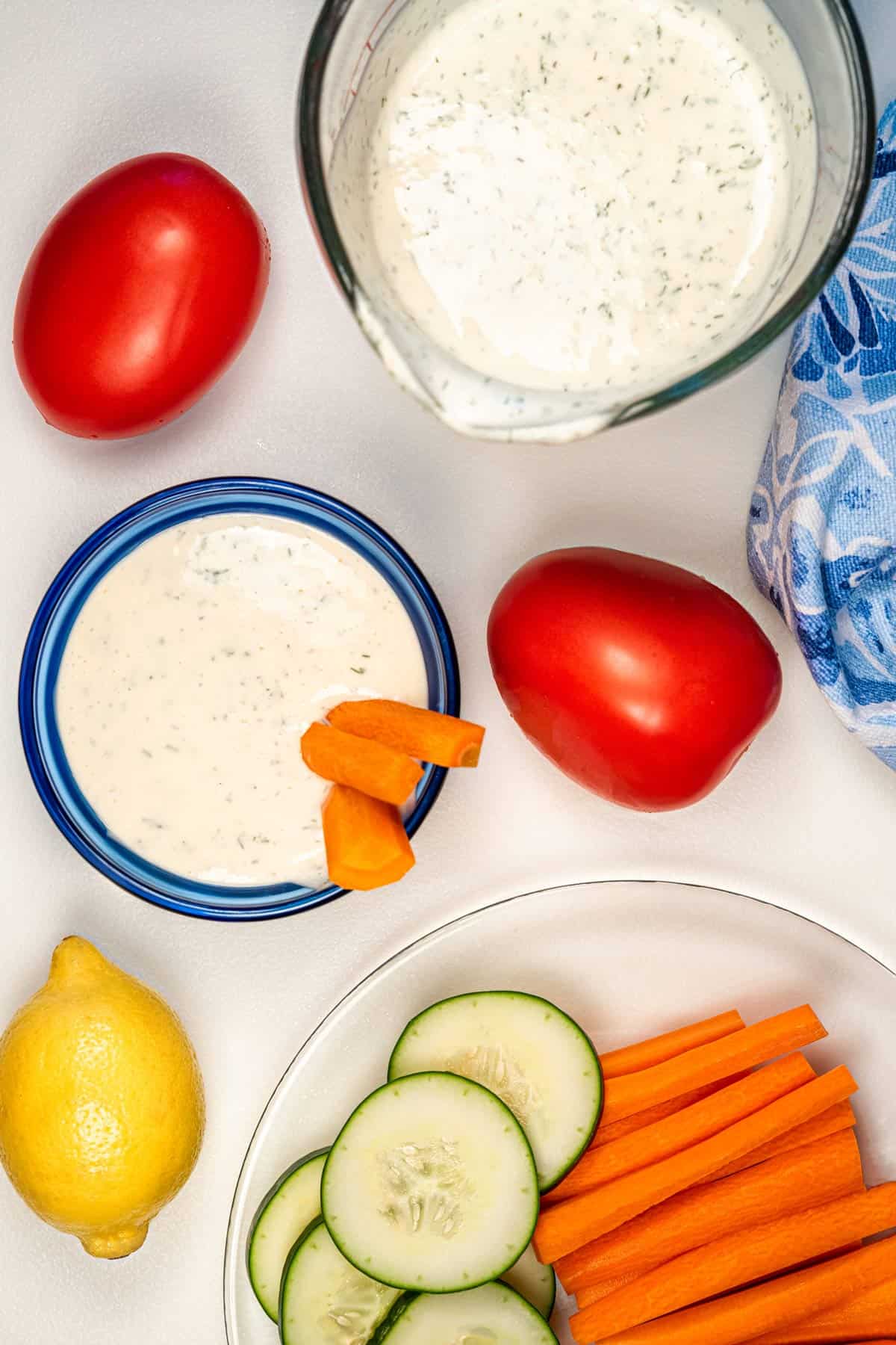 Dish of vegan ranch dressing with carrot sticks in it with other vegetables surrounding the dish like tomatoes, a lemon, and some cucumber slices.