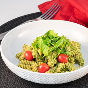 Bowl of vegan pesto pasta topped with arugula and tomatoes.