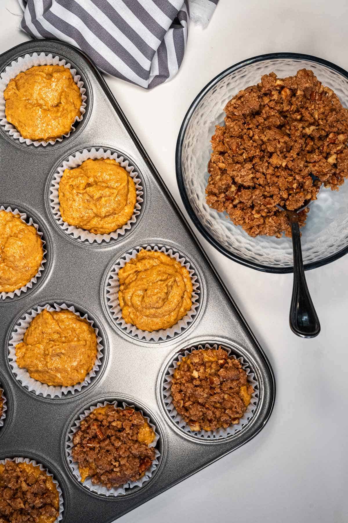 Unbaked Vegan Pumpkin Muffins in the tin ready to be topped with the bowl of crumble topping on the side.
