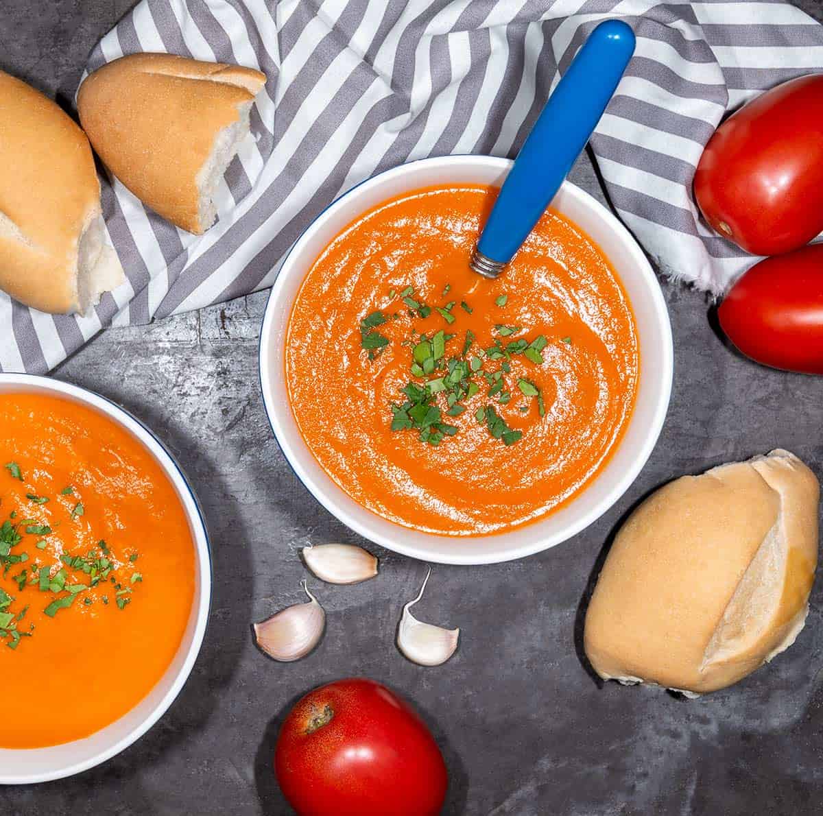 Overhead view of two bowls of vegan tomato soup with bread, garlic, and roma tomatoes in the background.