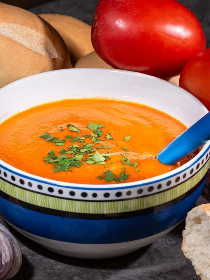 Bowl of vegan tomato soup with bread, garlic, and roma tomatoes in the background.