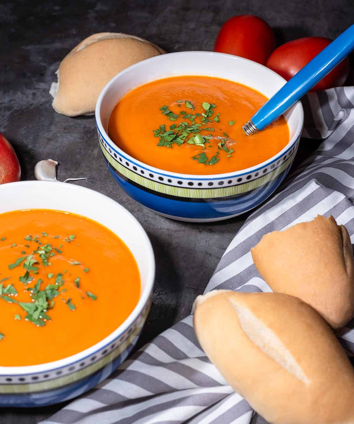 Two bowls of vegan tomato soup with bread and roma tomatoes in the background.