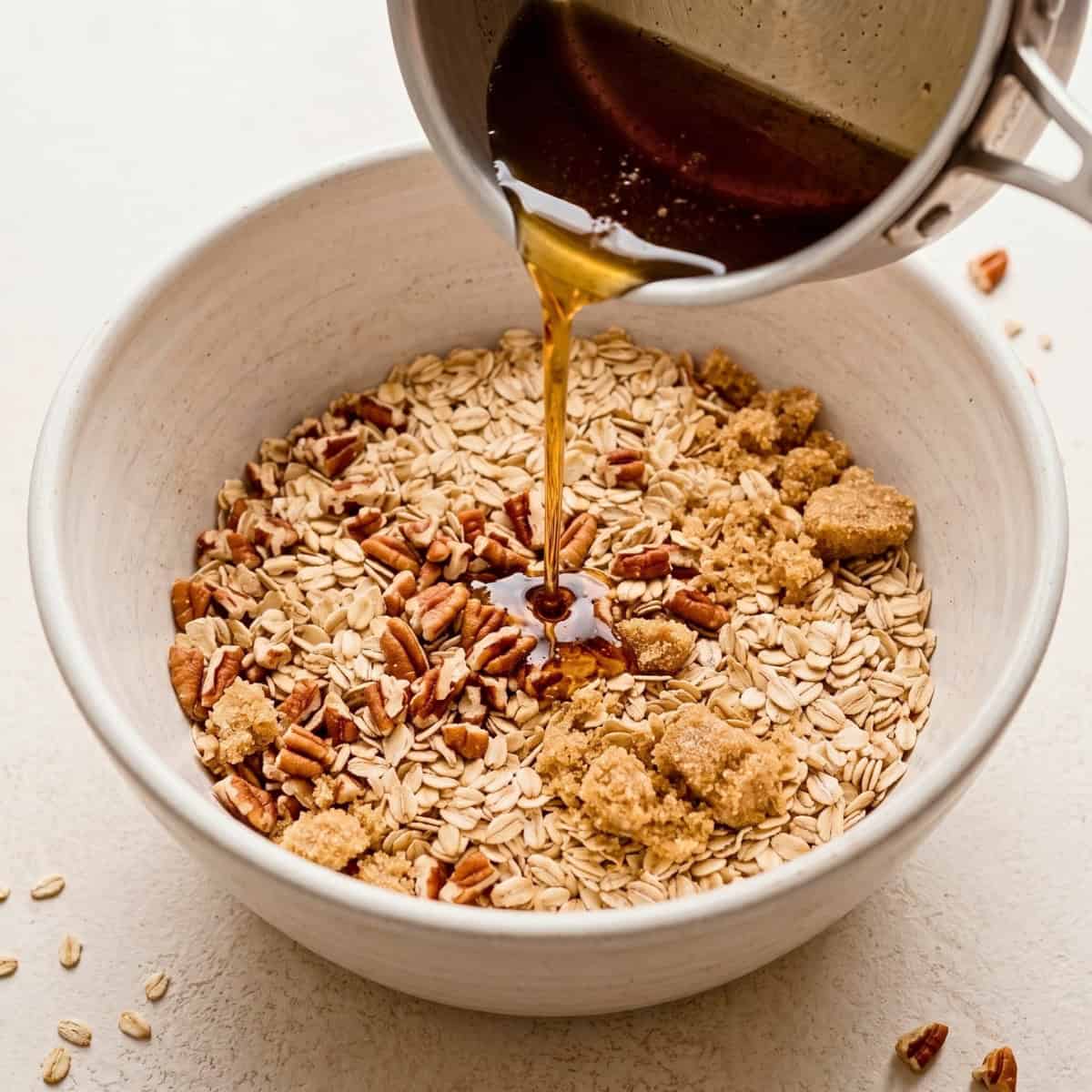 Pouring warm maple syrup and melted coconut oil over oats and chopped pecans in a mixing bowl.