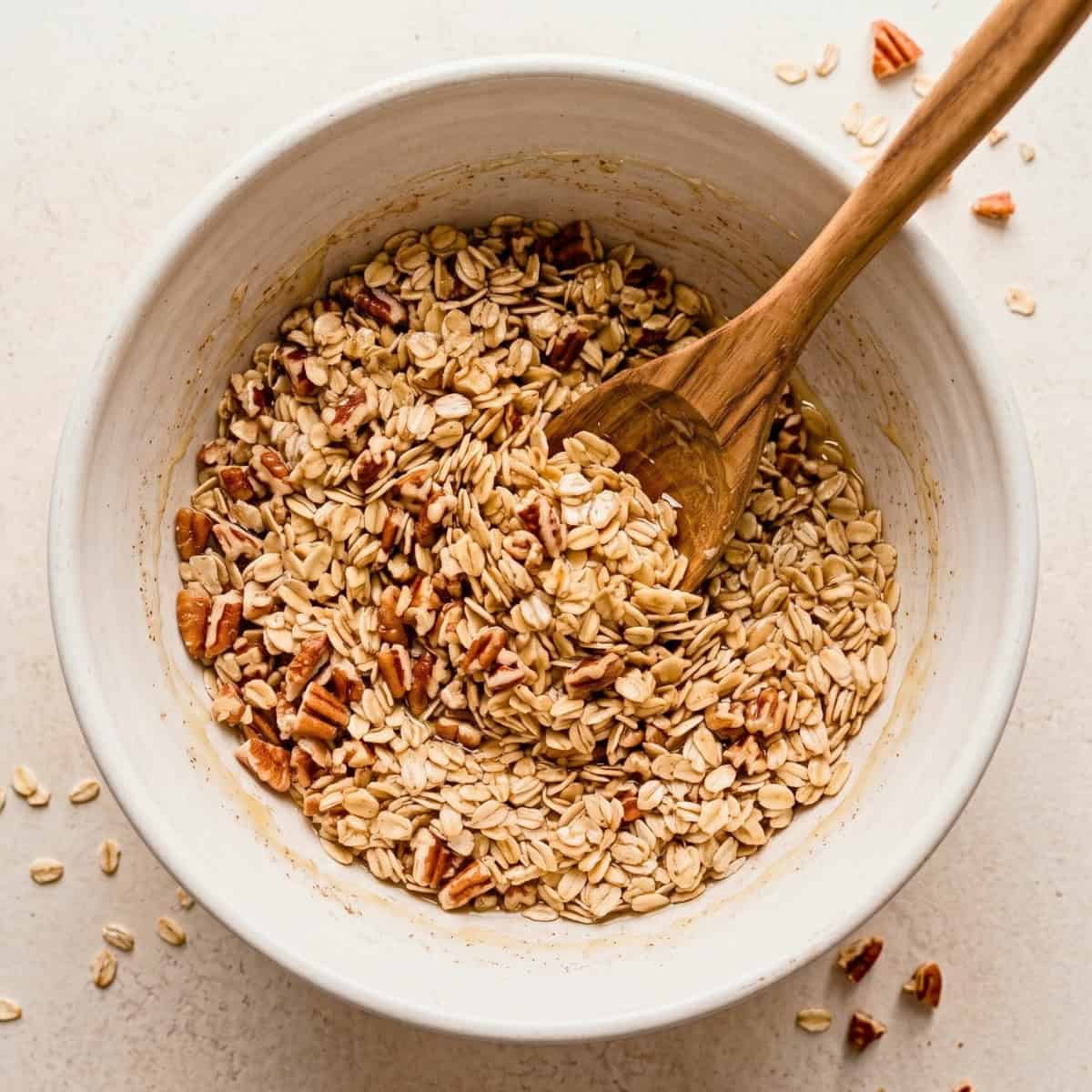 Mixing rolled oats, chopped pecans, and brown sugar in a white bowl with a wooden spoon.