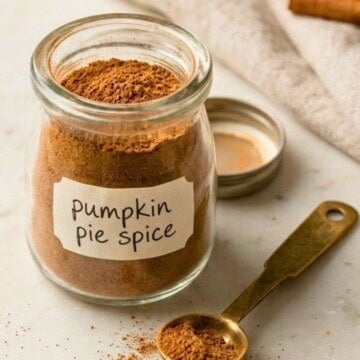 Close-up of homemade pumpkin pie spice in a labeled glass jar with a brass measuring spoon on a marble countertop.