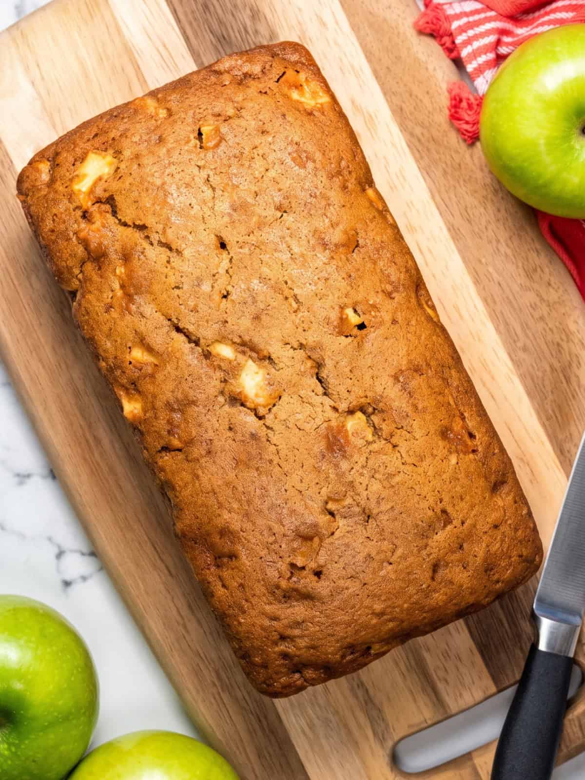 Overhead view of a golden brown vegan apple bread loaf on a wooden cutting board with fresh green apples.