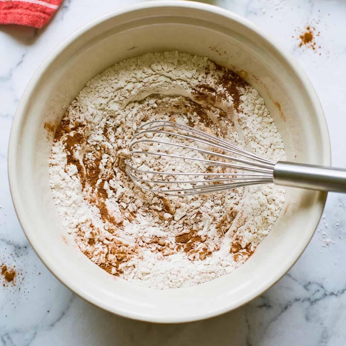 Flour, cinnamon, and other dry ingredients being mixed with a whisk in a bowl.
