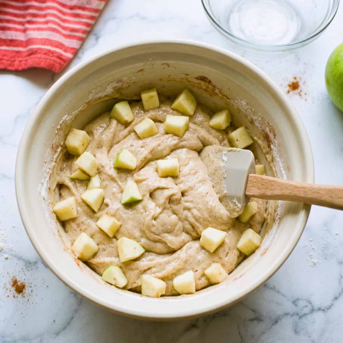 Vegan apple bread batter with chunks of fresh diced apples in a mixing bowl with a wooden spoon.