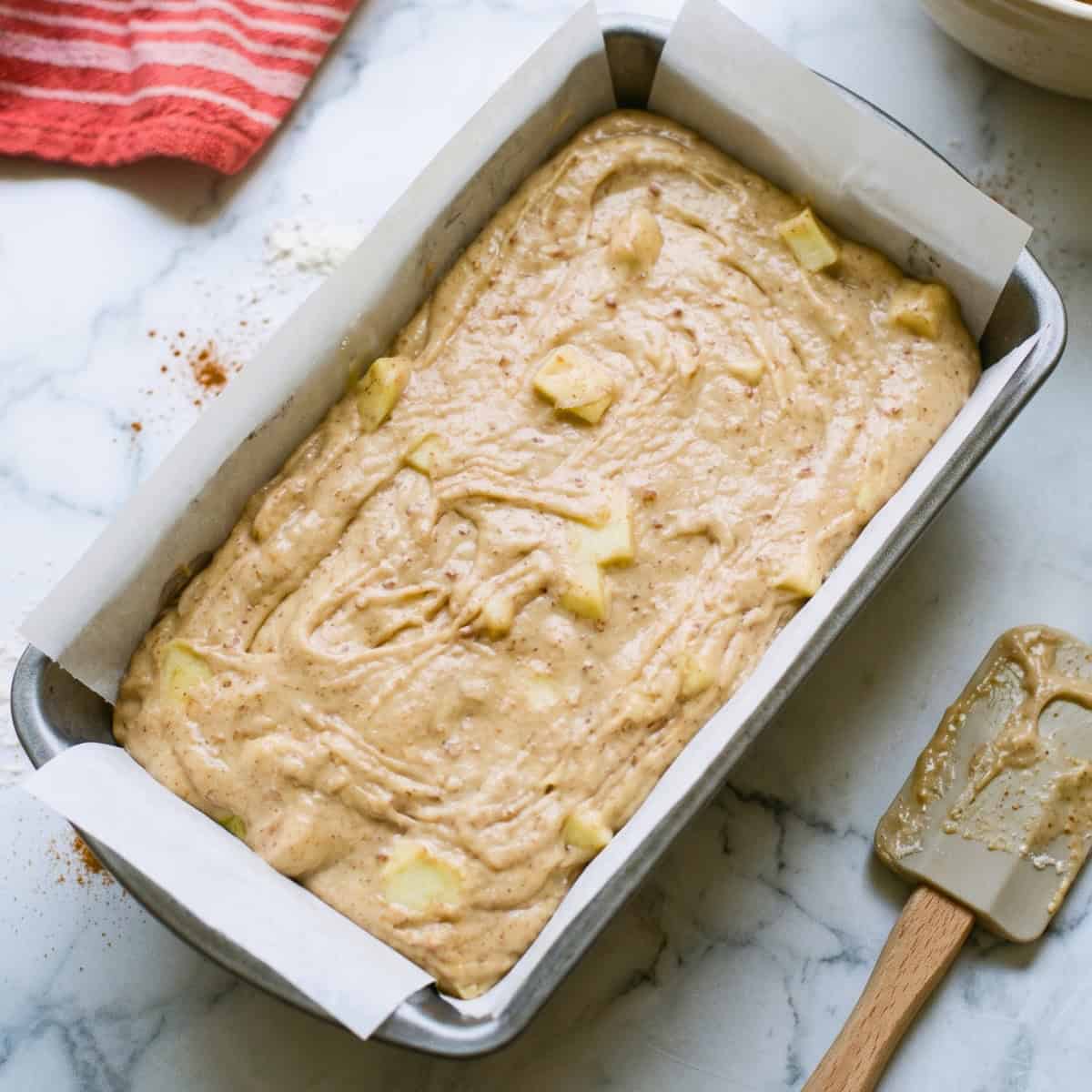 Unbaked vegan apple bread batter with diced apples in a parchment-lined loaf pan.
