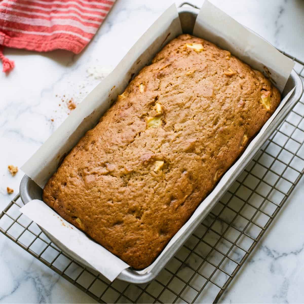 Freshly baked vegan apple bread with a golden brown crust cooling in a loaf pan on a wire rack.