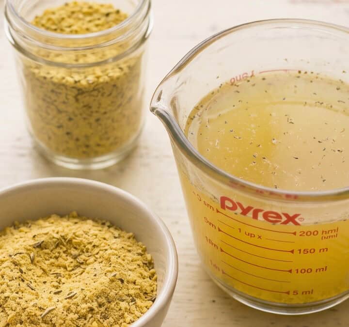 Close-up of homemade vegan chicken broth in a glass measuring cup beside a bowl and jar of bouillon powder.