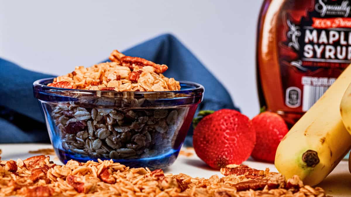 Homemade maple pecan granola in a blue glass dish with fruit and maple syrup in the background.