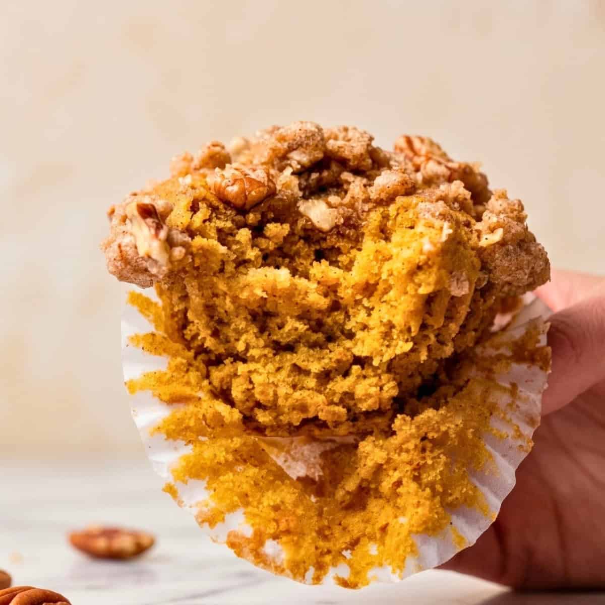 Someone holding up one baked vegan pumpkin muffin with pecan streusel and showing what the inside looks like.